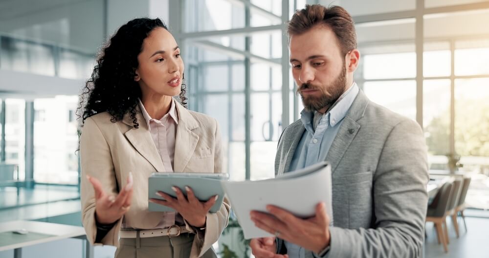 A woman and a man in professional suits discussing business documents and a tablet in a modern office setting.