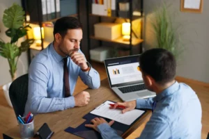 Two businessmen reviewing financial charts on a laptop and a paper contract at a wooden desk in an office.